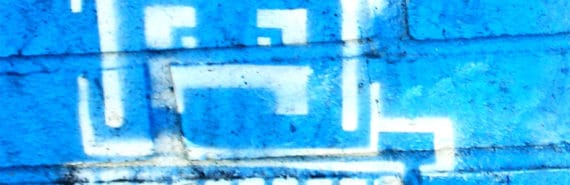 A computer with a frowning face is painted on a bright blue brick wall in white