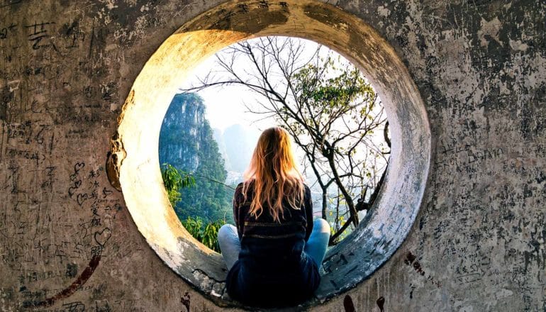 A woman sits in a hole in a concrete wall that looks out to a natural environment