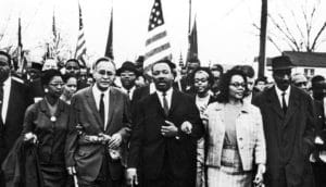 Martin Luther King Jr. walks at the center of a large marching crowd. American flags hang in the distance.