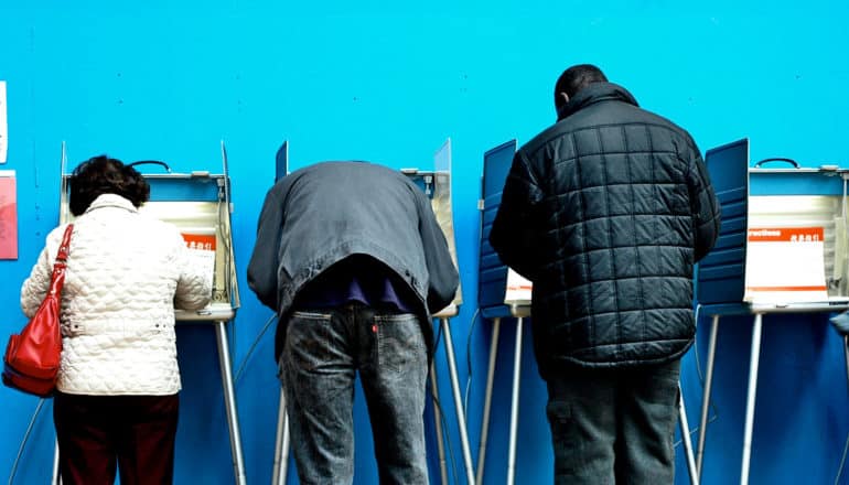 3 voters stand at voting booths filling out their ballots against a blue wall