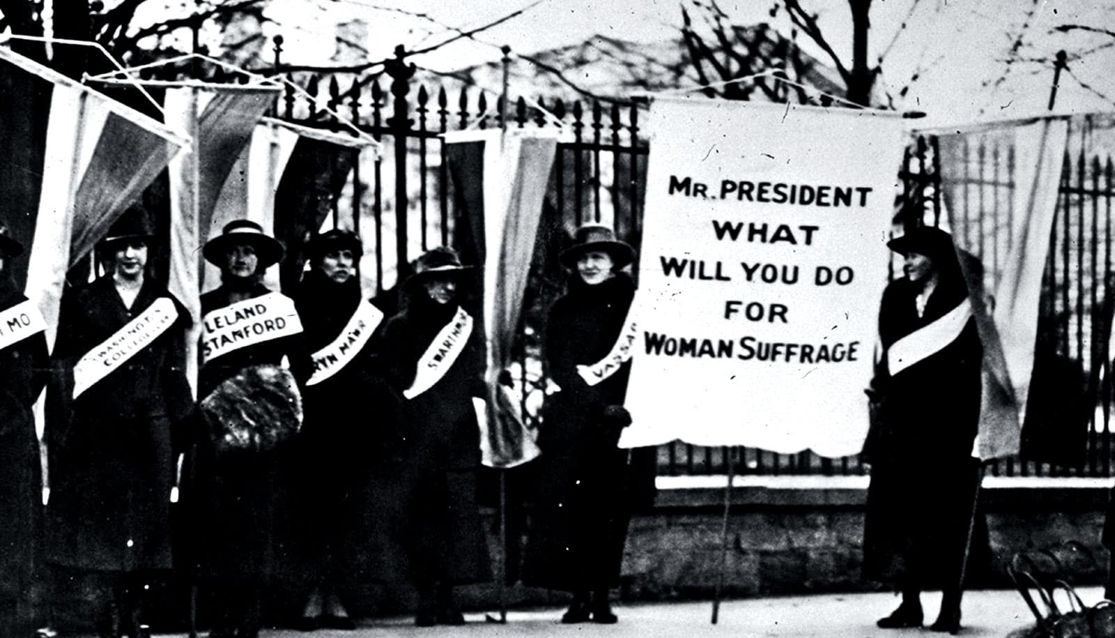 Women picket outside the White House, one holding a sign that reads, "Mr. President What Will You Do For Woman Suffrage"