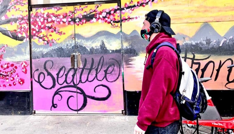 A man in a face mask walks past a wall covered in art and writing that reads "Seattle"