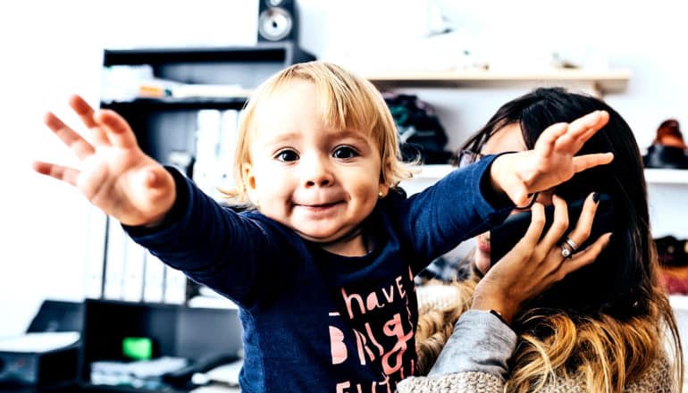 A baby girl reaches for the camera while her mother takes a work call