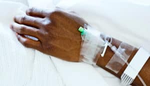 A man's hand lays on a hospital bed with various IV tubes in his arm