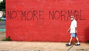An older man wearing a face mask walks past a red brick wall spraypainted with the phase, "No More Normal"
