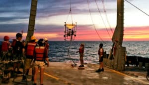 Researchers stand on the deck of a ship while lowering the device by winch into the water. The sun sets in the background.