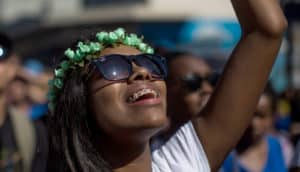 person in flower crown and sunglasses looks up and raises arm in praise