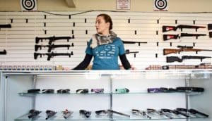 person stands behind case of handguns; rifles on wall behind