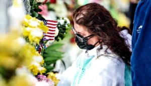 A woman looks down while near flowers and an American flag honoring shooting victims