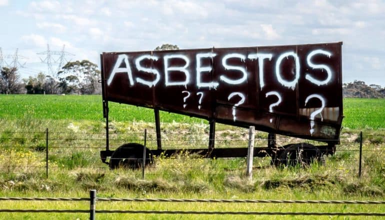 rusty sign in field says "ASBESTOS???" in white spray paint
