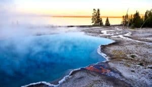 A blue hot spring has steam rising from it as the sun sets in the background.