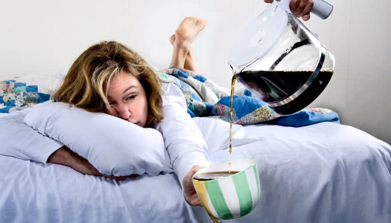 A woman waking up in bed holds a coffee cup as someone else pours coffee into her cup