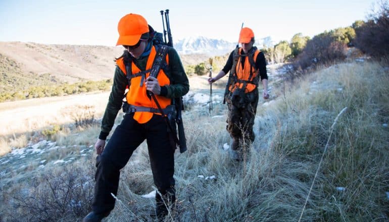 two people in neon orange gear with guns walk outdoors