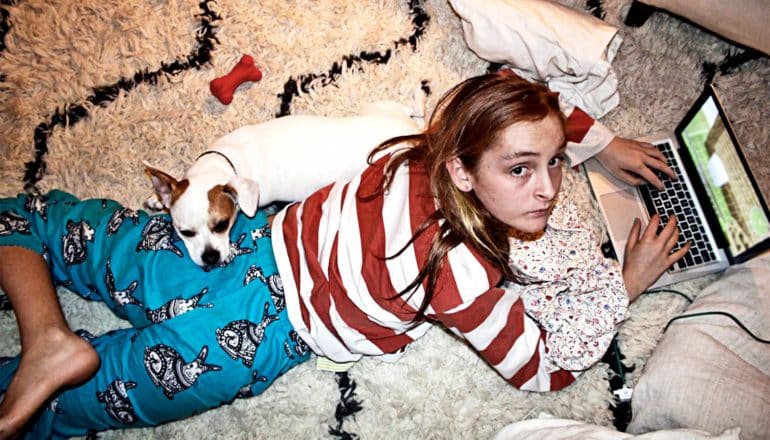 A young teen girl lays on her stomach on the floor while using a laptop and looking up at the camera