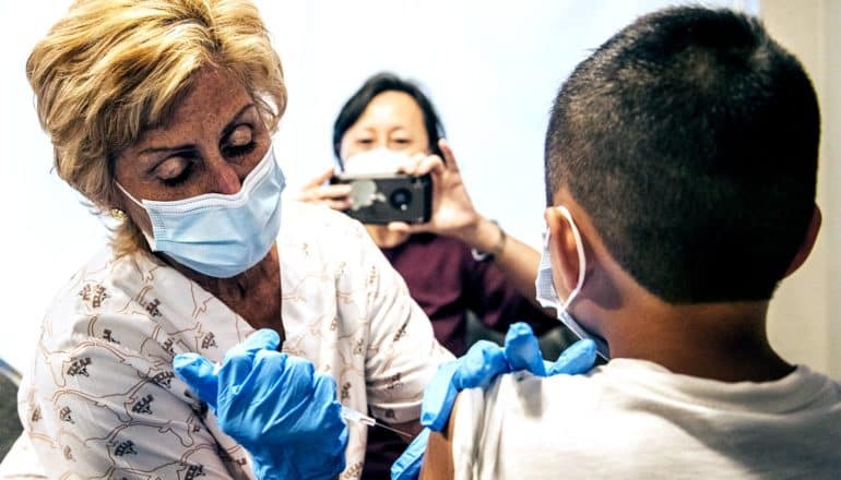 A nurse gives a child the COVID-19 vaccine as his mother takes a photo with her phone