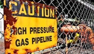 A yellow and black sign on a fence reads "Caution: High Pressure Gas Pipeline"