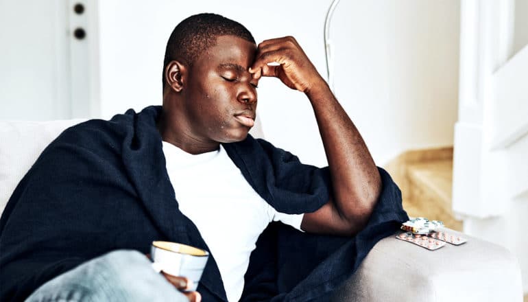 A man raises a hand to his head while wearing a blanket around his shoulders and holding a cup of tea