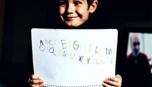 A young boy holds up a sheet of paper with letters written in different colors while smiling