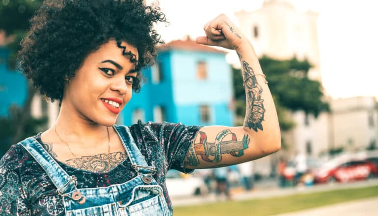 smiling, tattooed person flexes one arm outdoors