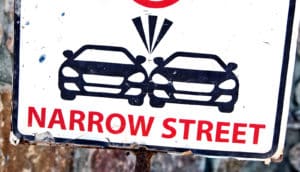 A sign reads "Narrow Street" with two cars scraping each other above
