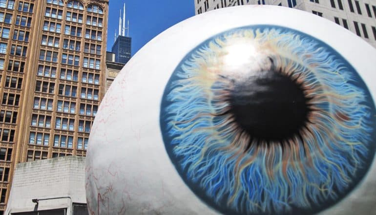 A giant statue of an eyeball sits in front of skyscrapers and a blue sky