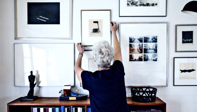 Een oudere vrouw hangt een ingelijste foto aan een muur vol andere ingelijste kunst