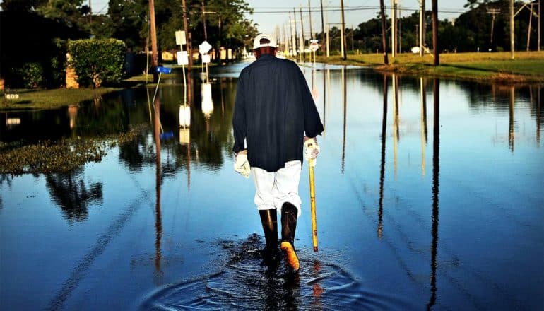A man walks with a cane and rain boots through a flooded residential street