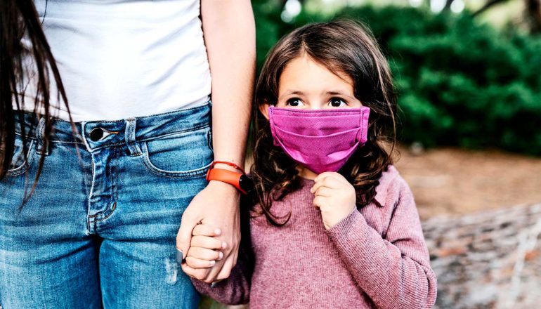 A mother holds a young girl's hand as she walks wearing a purple face mask