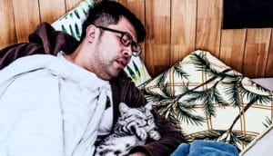 A man naps under a blanket on some pillows against a wood-paneled wall while holding a small cat
