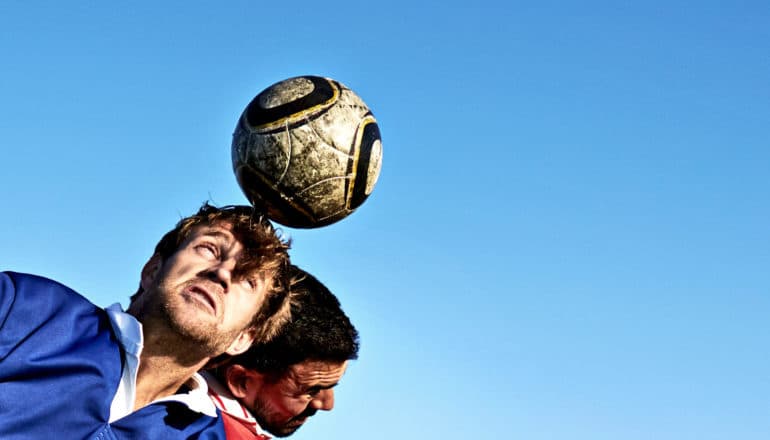 Two soccer players, one in red and one in blue, both leap to head the ball against a blue sky