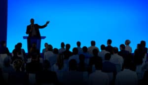 silhouette scene of person speaking at podium before seated crowd