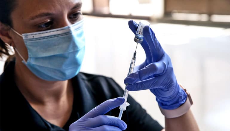 A health care worker fills a syringe with the COVID-19 vaccine while wearing a face mask and purple gloves