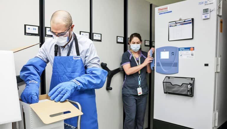 person opens box with large safety gloves; another waits to open freezer door