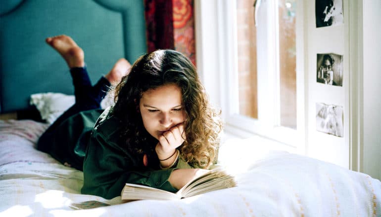 A teen girl lays on her bed reading a book