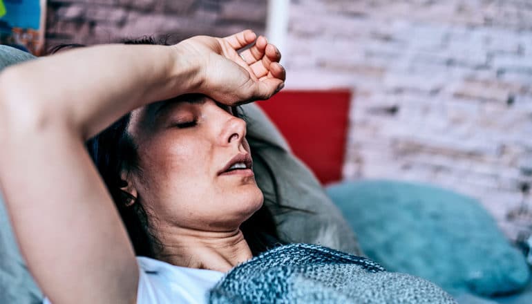 A woman laying in bed puts her arm over her forehead and has a pained look on her face