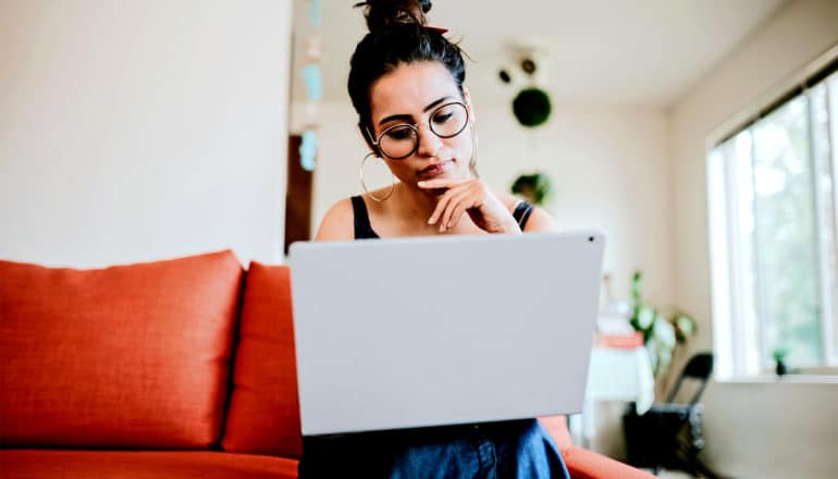 A young woman touches her chin while looking down at her laptop
