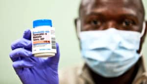Imbula holds up a bottle of fluvoxamine while wearing purple gloves and a blue medical mask