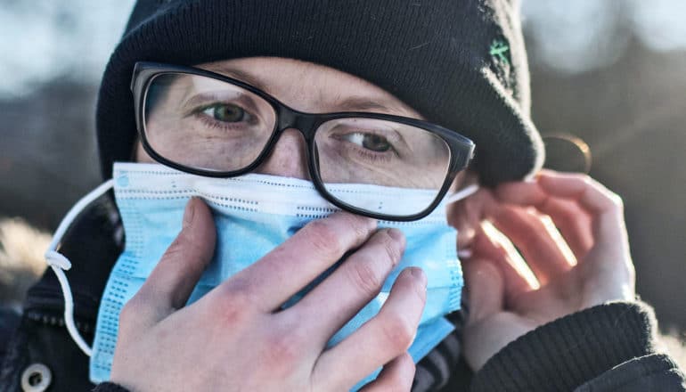 A person puts on their medical mask while wearing a winter hat, coat, and glasses