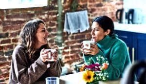 Two women sit at an office kitchen table sharing tea and talking
