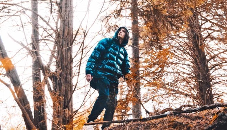 A young man in a blue puffer jacket walks through a forest during a rain shower