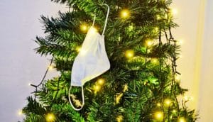 A medical face mask hangs from a green Christmas tree covered in yellow lights