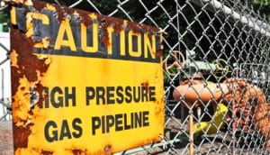 A yellow, rusting sign reads "Caution: High pressure gas pipeline"