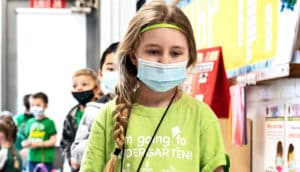 A young girl wearing a medical mask stands at the front of a line of young school kids in a classroom