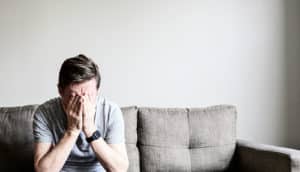 A man sits alone on a couch covering his face with his hands