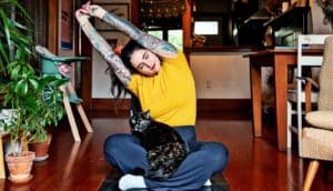A young woman stretches while sitting on a yoga mat in her apartment. Her cat sits on her lap.