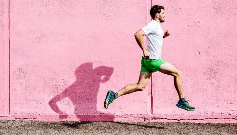 A man in a white t-shirt and green running shorts runs in front of a pink wall