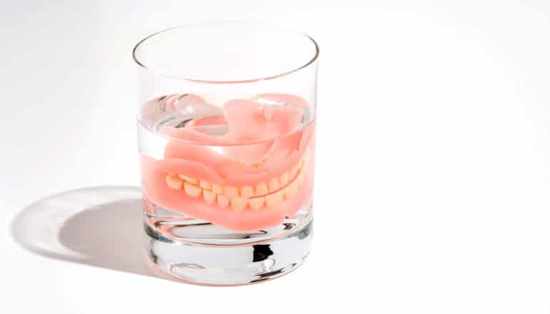 Dentures sit in a glass of water against a white background