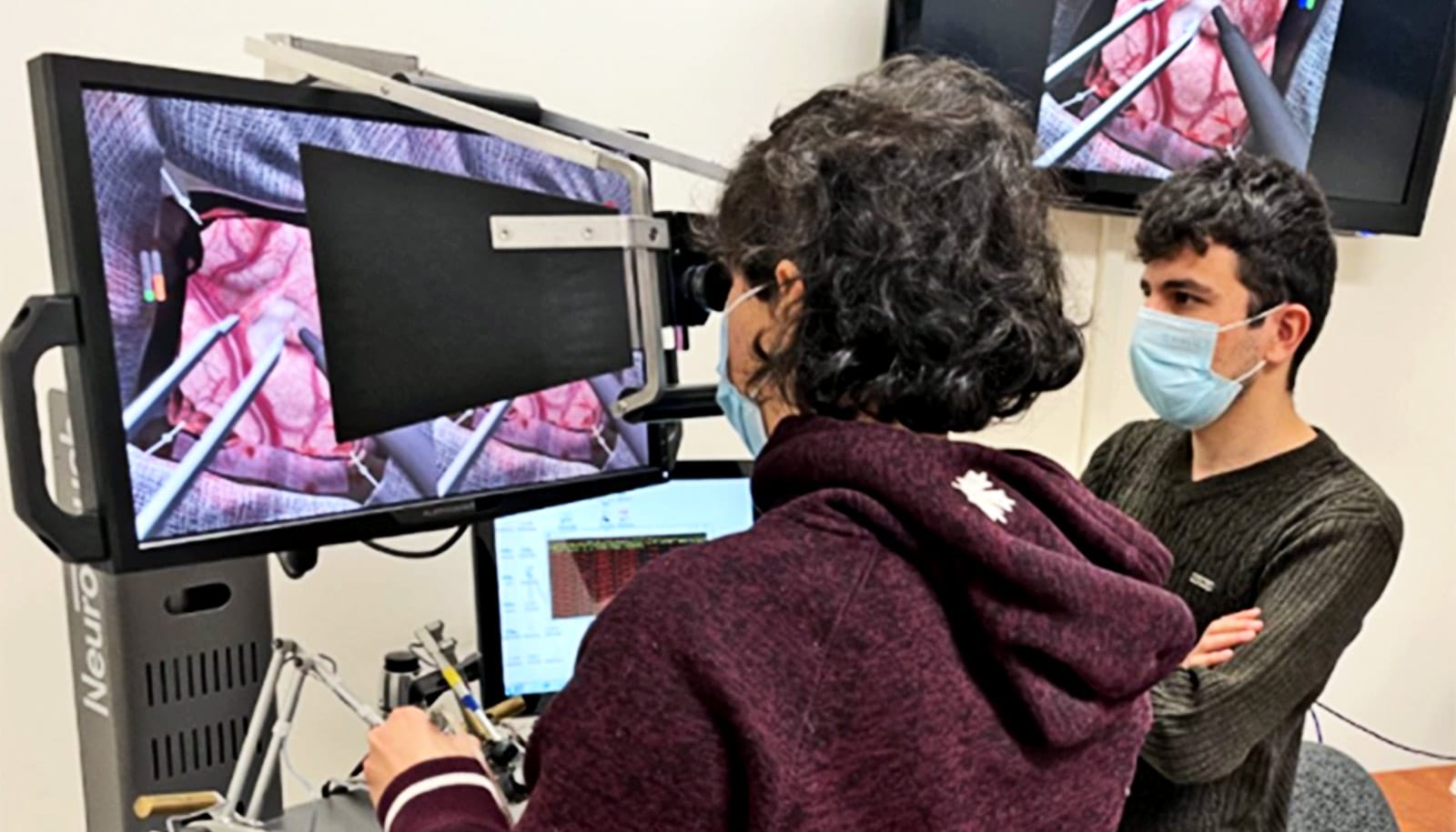 A young woman looks into a scope while operating a surgical robot. Virtual simulation of brain surgery appears on screens around her.