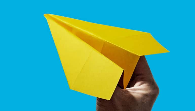 Een persoon houdt een geel papieren vliegtuigje tegen een blauwe achtergrond