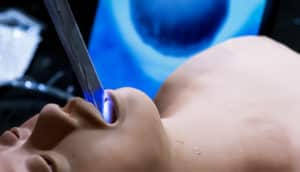 The intubation device is in the mouth of a surgical dummy, with light shining on the inside of the mouth and a screen showing a close-up view on the monitor in the background.